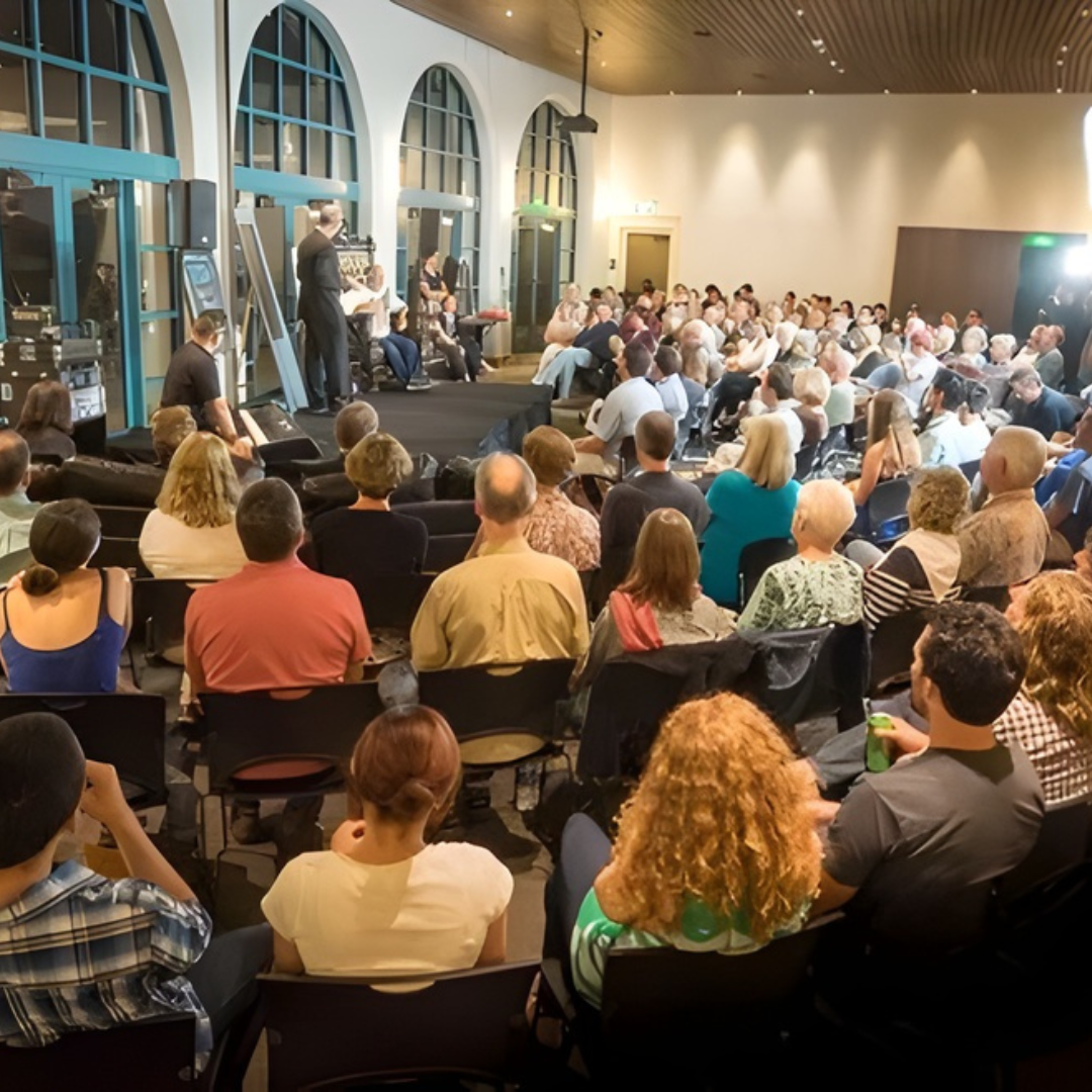 Large audience seated in a bright hall watching a stage presentation near tall arched windows