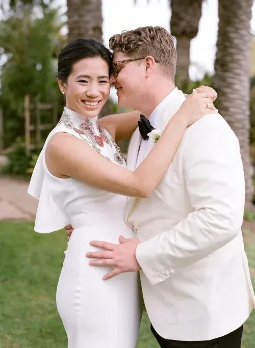 Two people in white wedding attire smiling and embracing outdoors by palm trees.