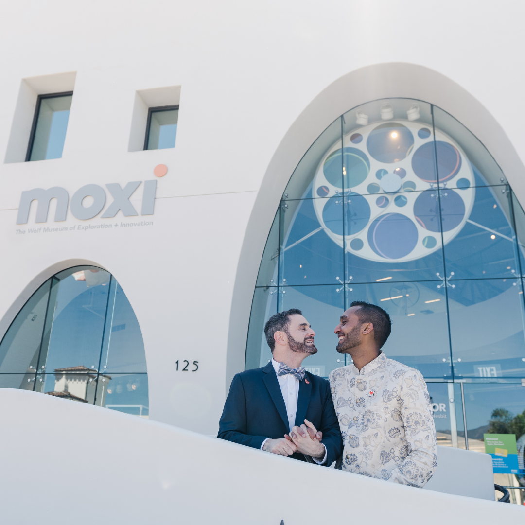 Two men in formal wear standing outside a white building with a large arched glass entrance and “moxi” sign