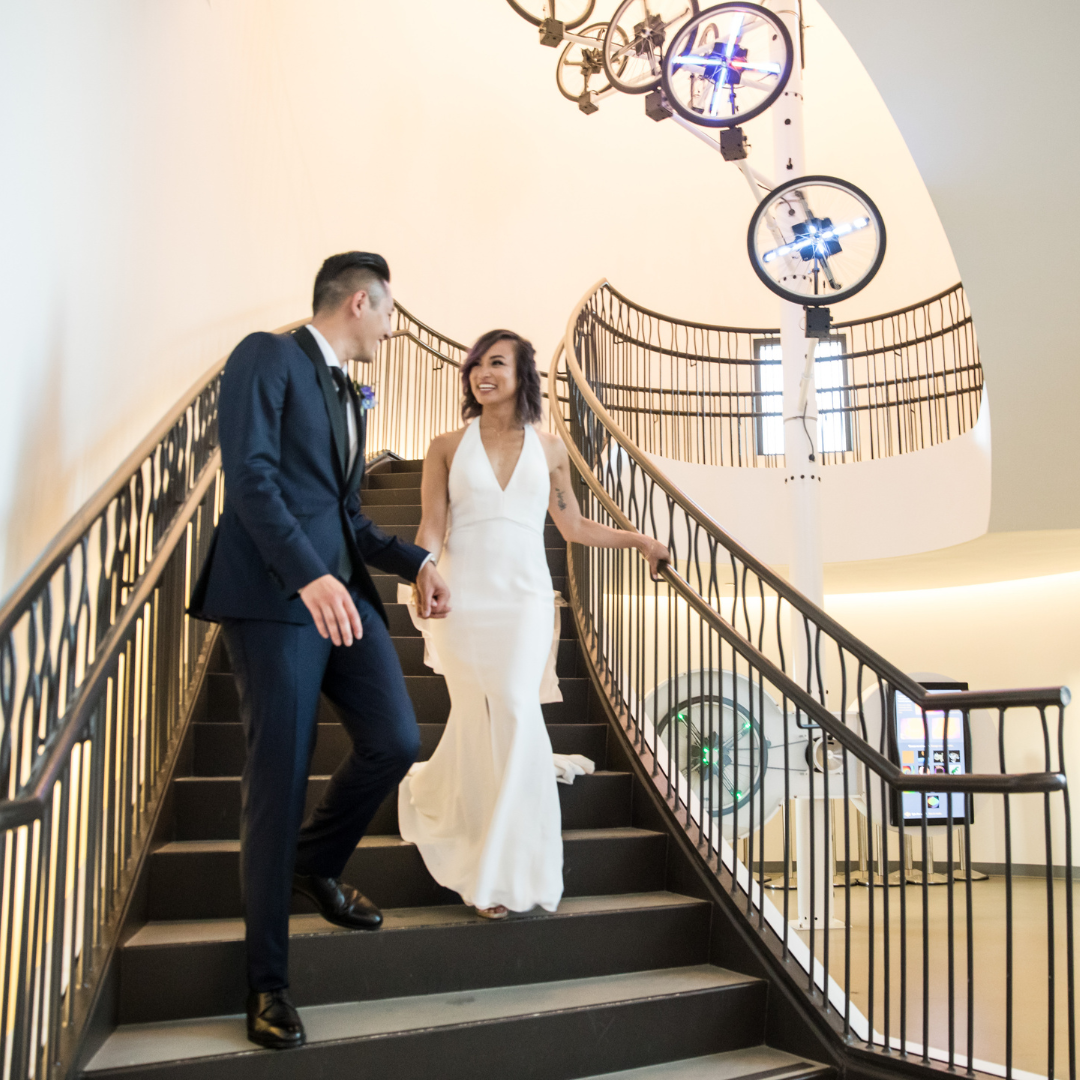 Couple in formal wear descending an ornate staircase in a bright interior.