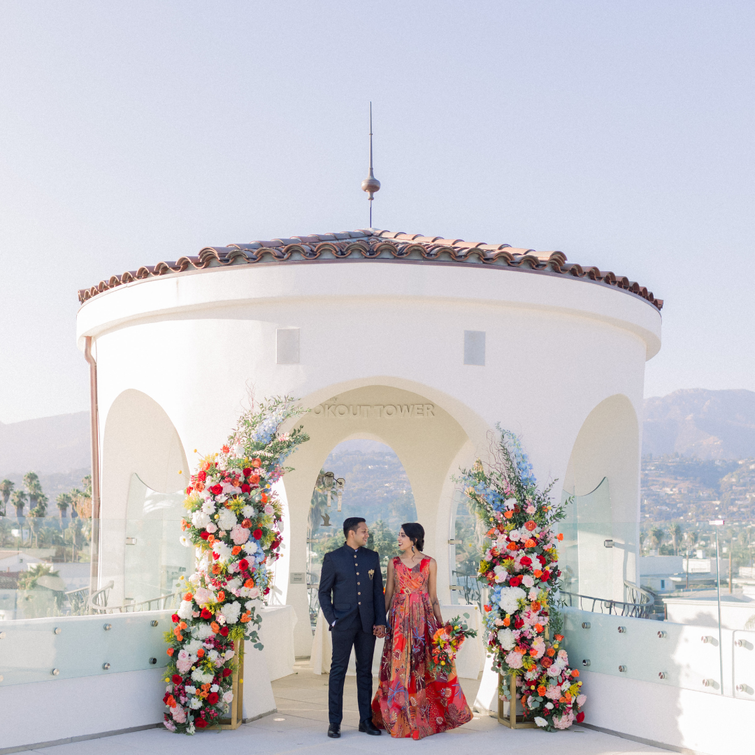 Couple posing under floral arch at a white rooftop venue with mountains in the background