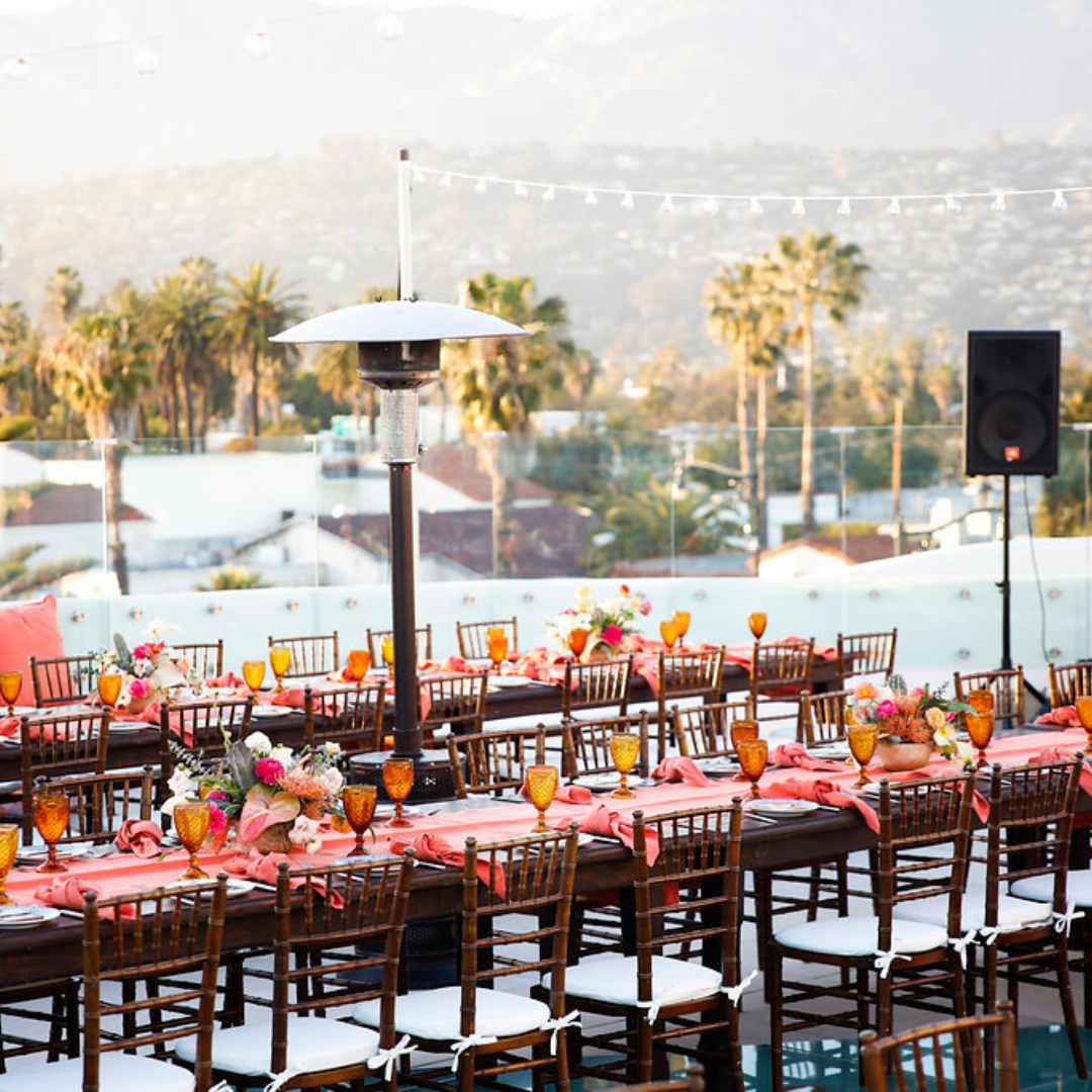Elegant outdoor dining setup with orange table settings, wooden chairs, palm trees, and a poolside view at sunset