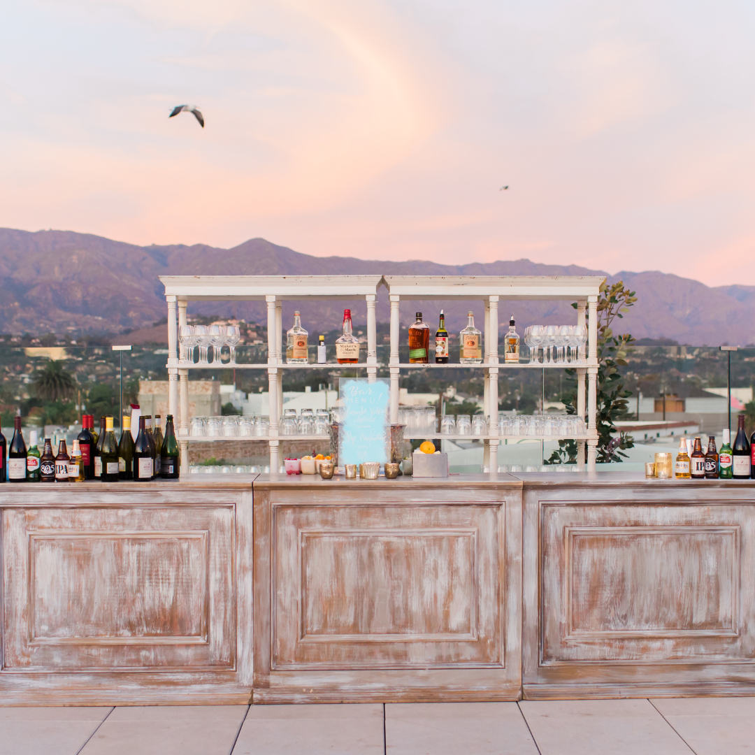 Bar with bottles and glasses on a wooden counter, with mountains and birds at sunset in the background