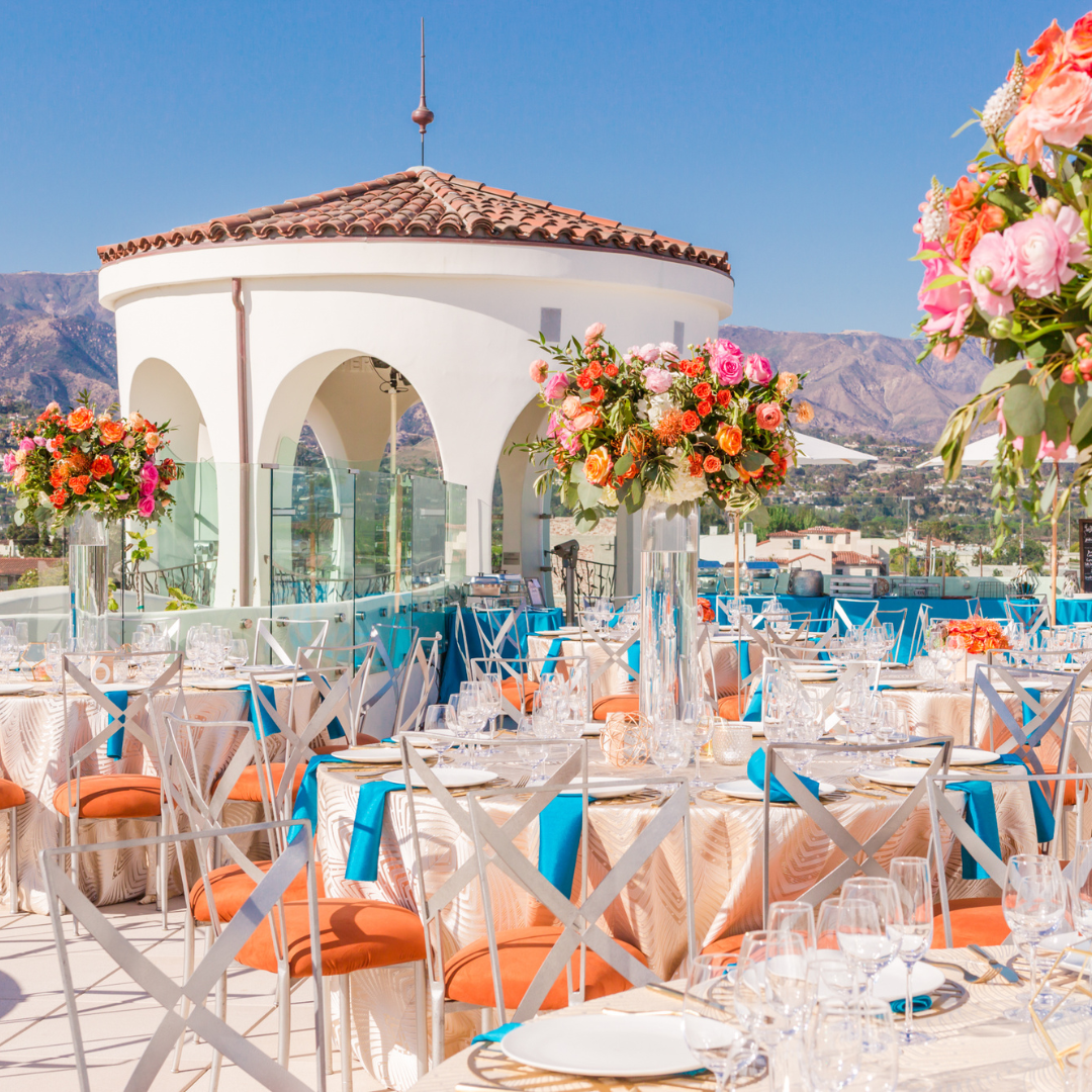 Elegant outdoor wedding reception with floral centerpieces, white chairs, and blue accents by a seaside terrace