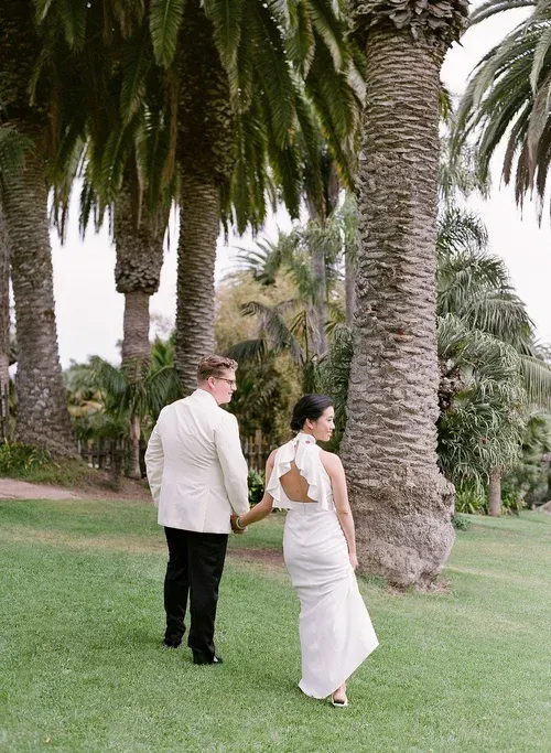 Couple walking hand in hand on a grassy path among tall palm trees at sunset