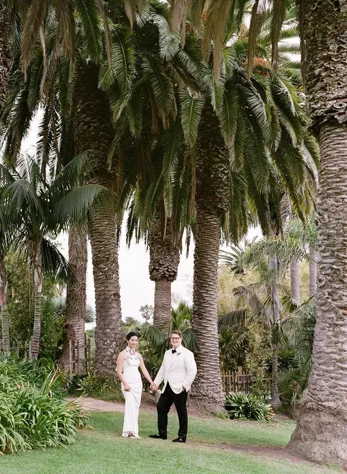 Couple holding hands in a lush palm garden, surrounded by tall trees and greenery.
