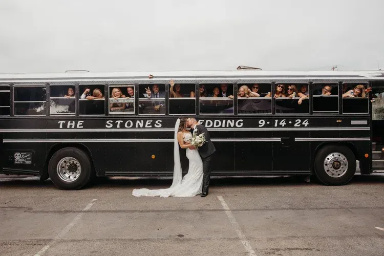 Bride and groom pose beside a black wedding bus labeled “The Stones” at a stone wall venue.