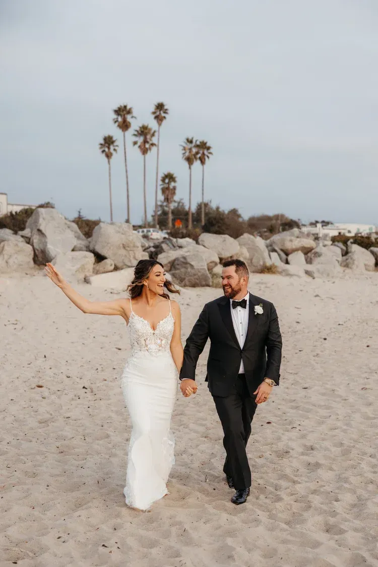 Bride and groom walking on a beach, bride holding up her dress with palm trees in the background