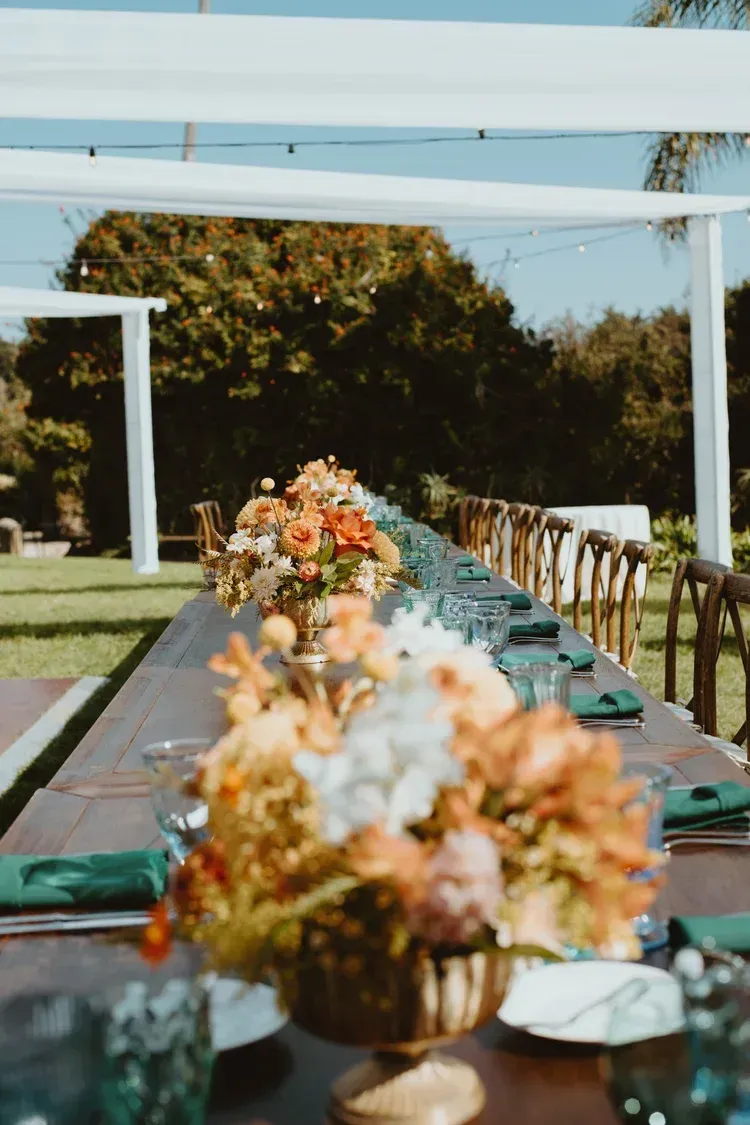 Long outdoor dining table with floral centerpieces under white pergola beams