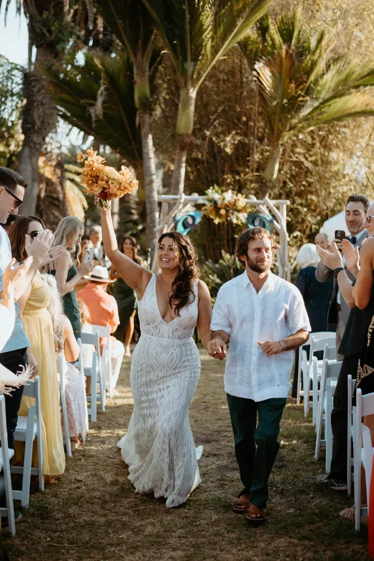 Newlyweds walking down a palm-lined aisle, guests cheering and taking photos outdoors