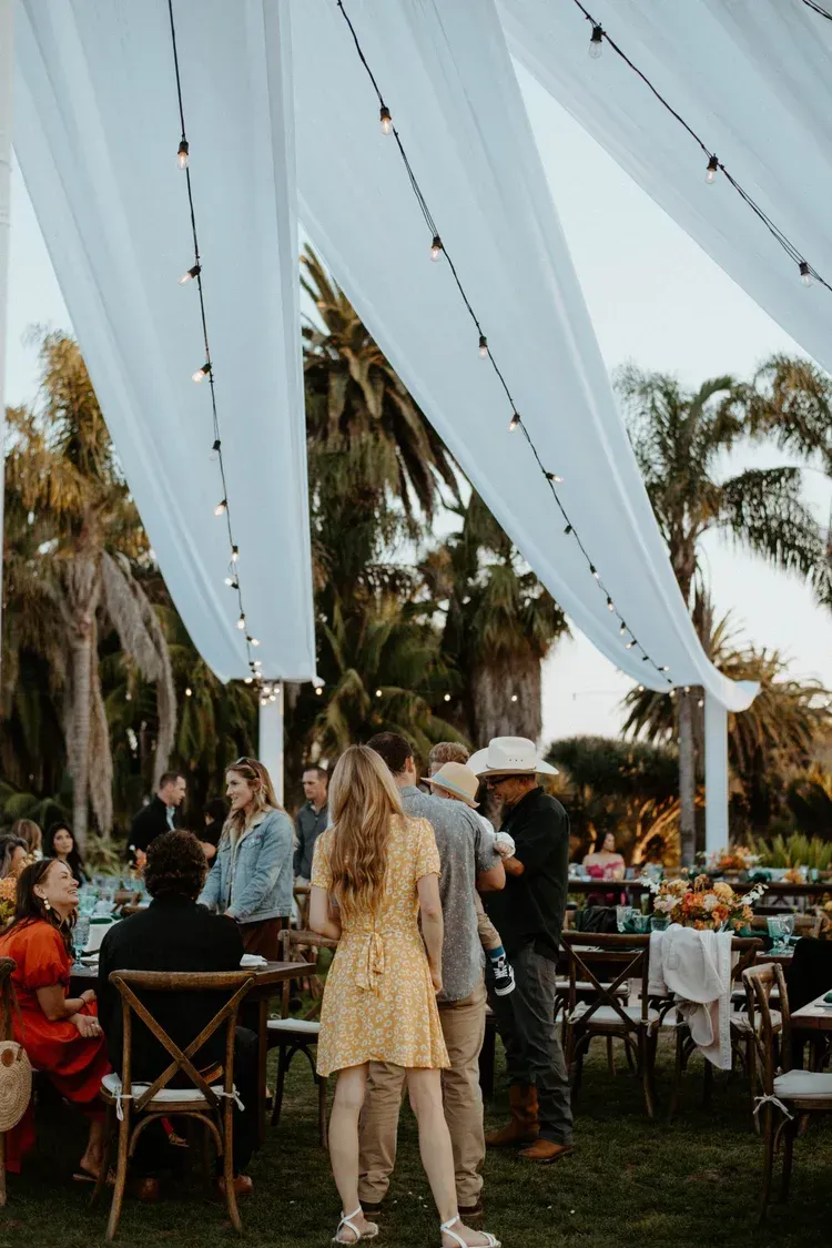 Guests mingle beneath white drapes at an outdoor garden reception with palm trees and tables.