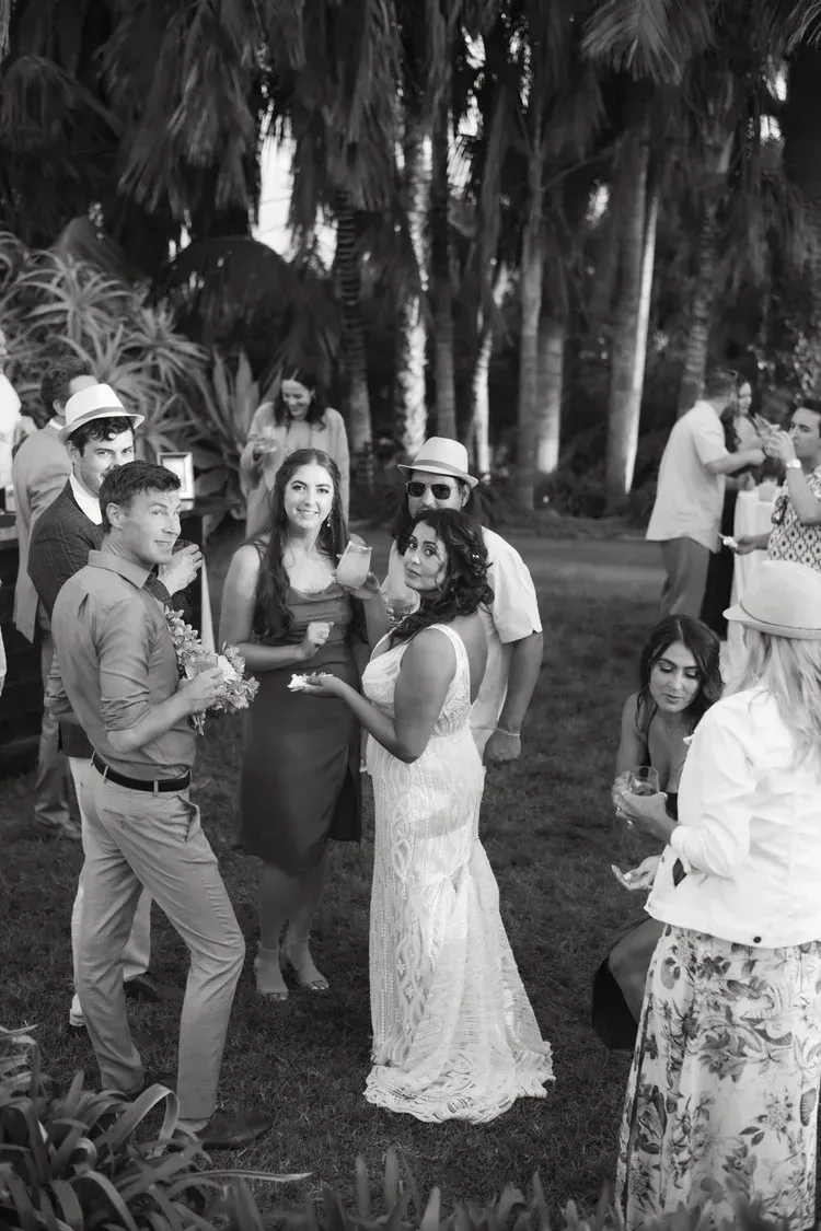 Outdoor group gathering with a woman in a white dress holding a cake, surrounded by smiling guests.