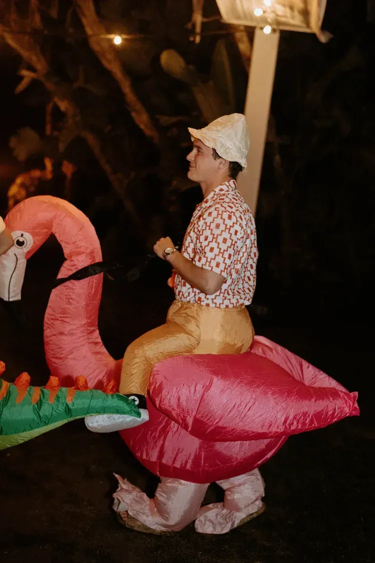 Person riding a pink flamingo float at night, wearing a white cap and patterned shirt.