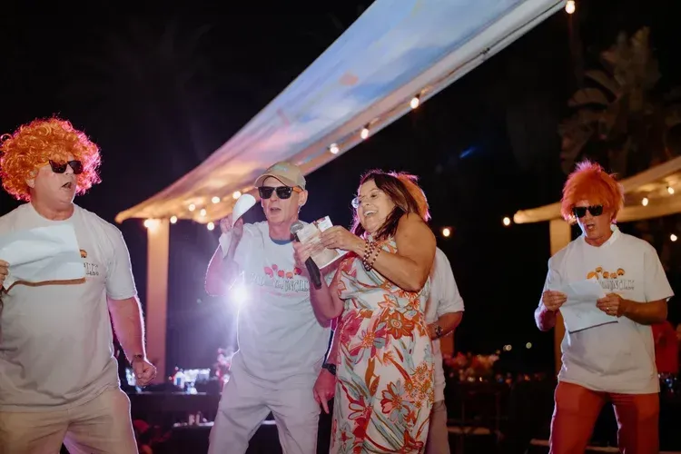 Four people dancing outdoors at night under string lights, wearing white outfits and colorful wigs.