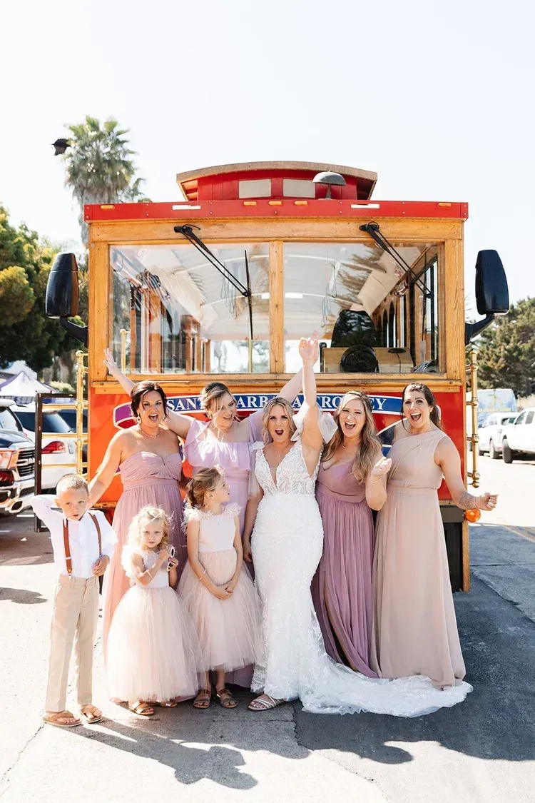 Group posing in front of a bright orange tram at an outdoor celebration, many in pastel formal dresses.