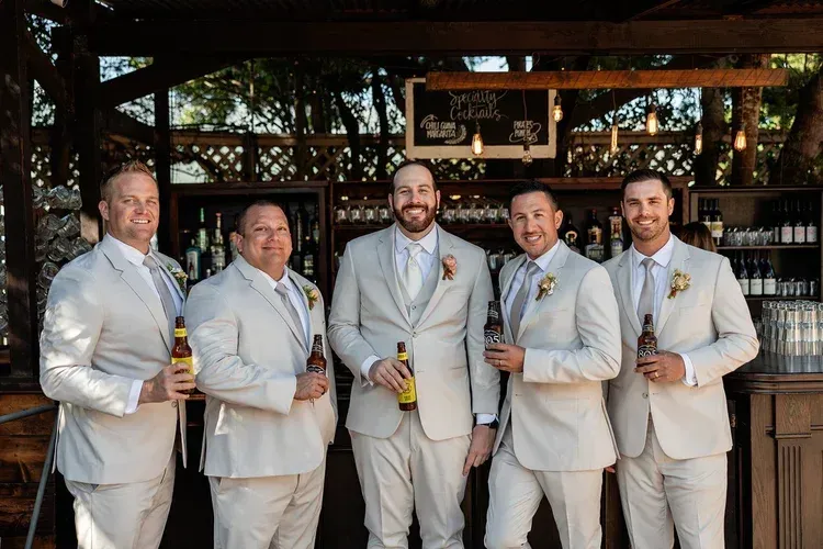 Five men in light gray suits holding drinks pose at a rustic bar, smiling indoors.