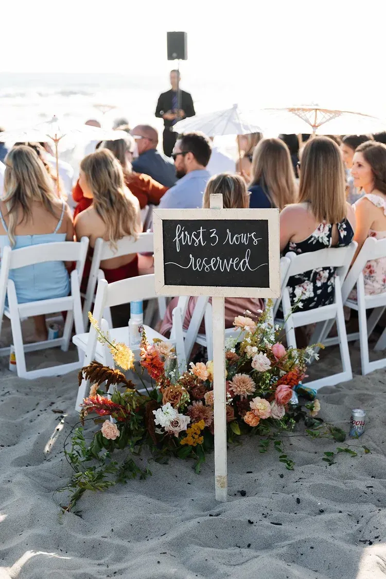 Wedding ceremony on the beach with seated guests, floral arrangement, and a chalkboard sign in the sand
