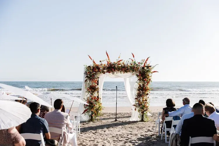 Beach wedding ceremony setup with floral arch facing the ocean and guests seated on the sand