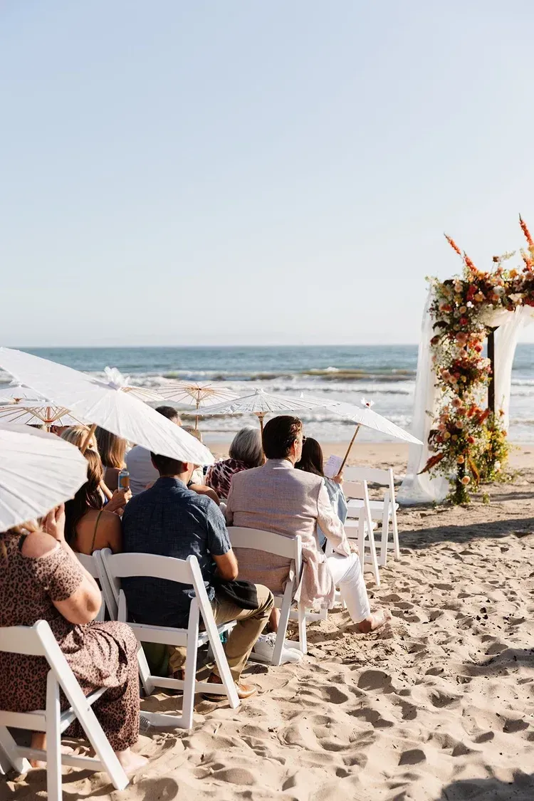 Beach wedding ceremony with seated guests facing a flower-decorated arch by the ocean