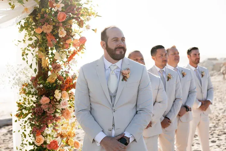 Groom and groomsmen in light suits stand beside a floral wedding arch outdoors.