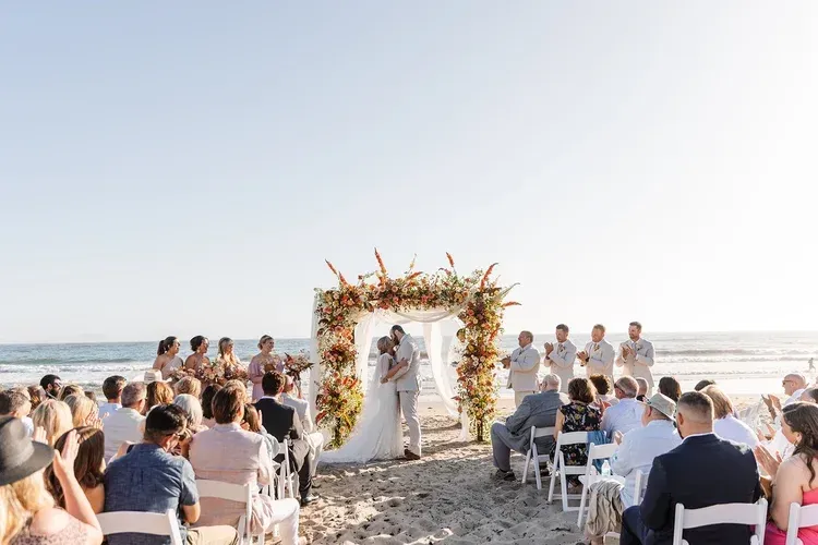Beach wedding ceremony under a floral arch with guests seated in the sand at sunset