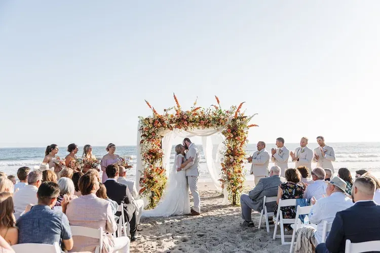 Beach wedding ceremony under a floral arch, with bride and groom exchanging vows as guests watch.