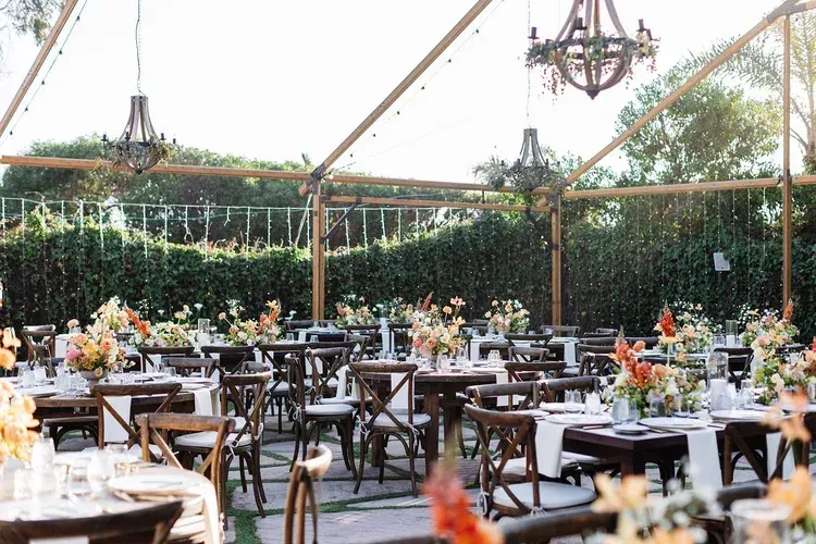 Outdoor event tent with black tables, white chairs, floral centerpieces, and string lights under a clear canopy