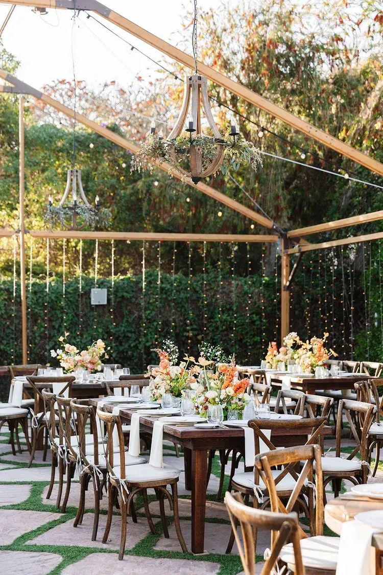 Outdoor patio reception with wooden tables, chairs, and string lights under a canopy