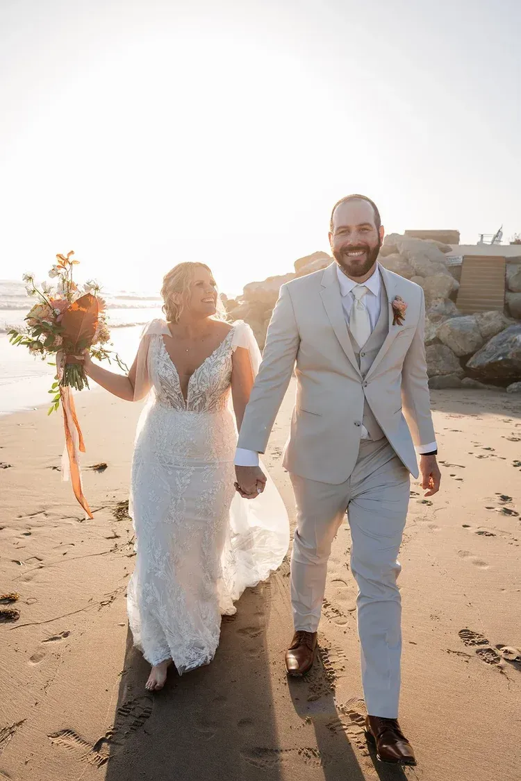 Bride and groom walking on a sandy beach at sunset, holding hands and smiling