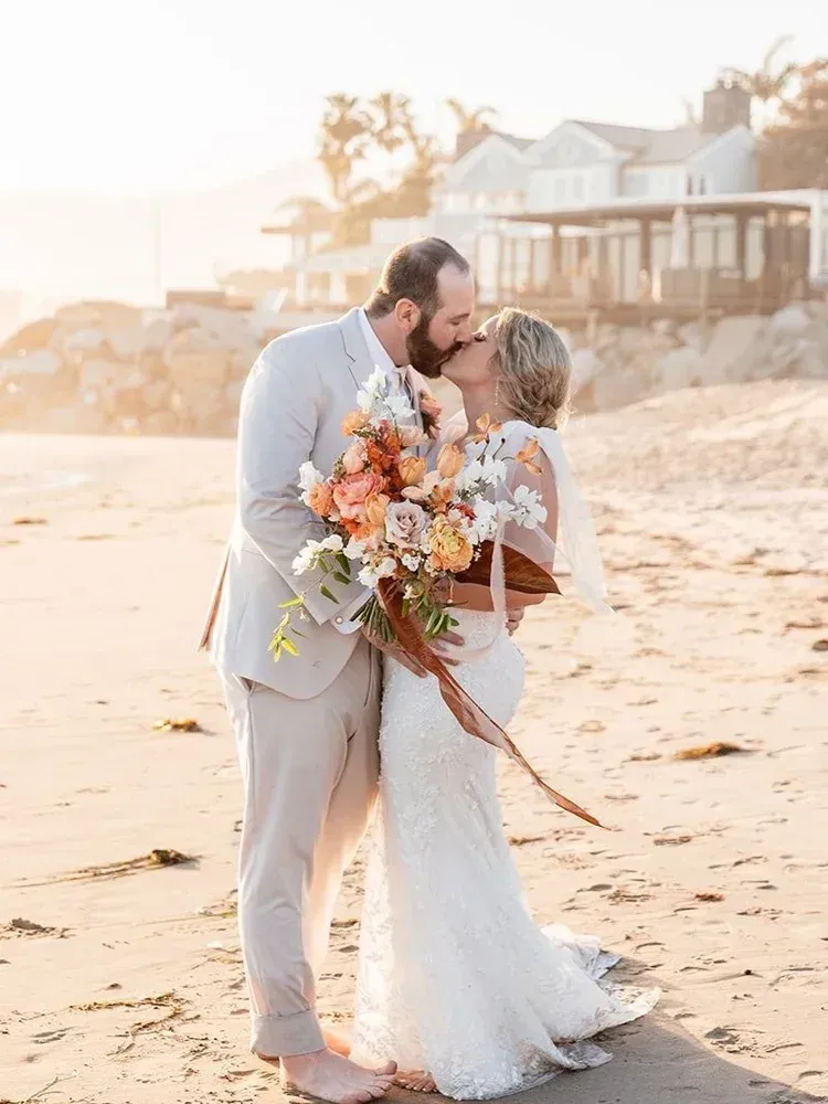 Couple kissing on a beach at sunset, bride holding a colorful bouquet, with oceanfront houses in the background