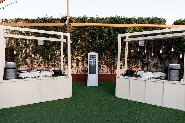 Outdoor buffet setup with white counters, wooden canopy, and green hedge backdrop