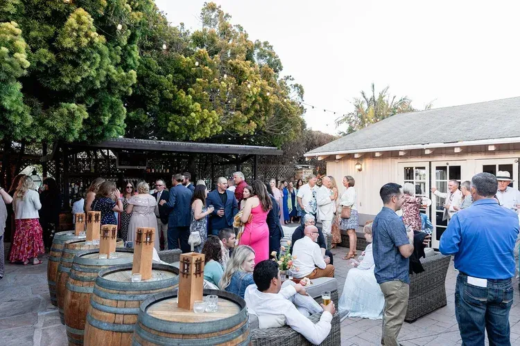 Guests mingling outdoors beside wooden barrels and a pavilion at a social gathering