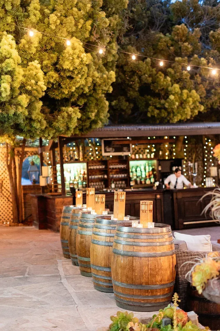 Outdoor bar with wooden barrels and string lights under leafy trees