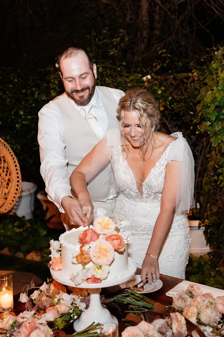 Bride and groom cutting a white wedding cake with peach flowers at an outdoor evening reception