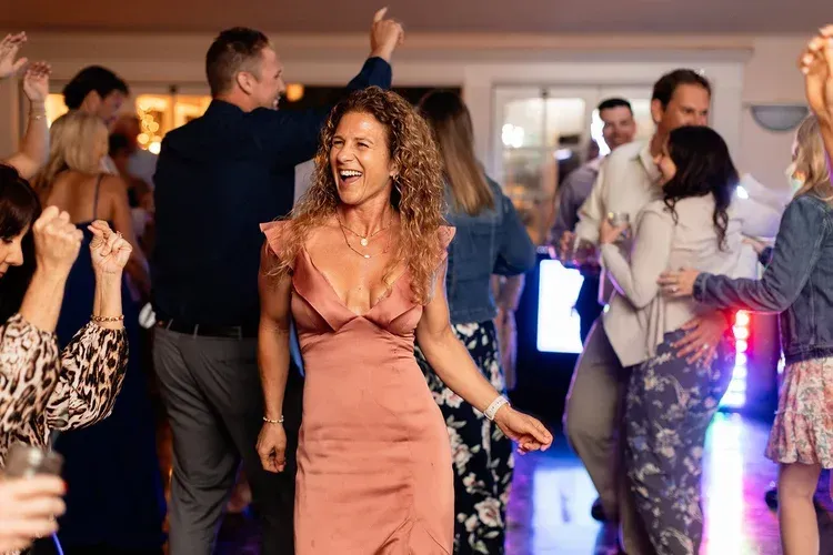 Guests dancing at a lively indoor party, with a woman in a pink dress smiling in the center.