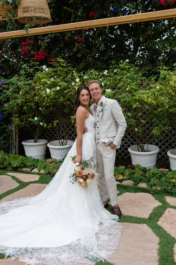 Bride and groom in white wedding attire posing in a garden with greenery and flowers.