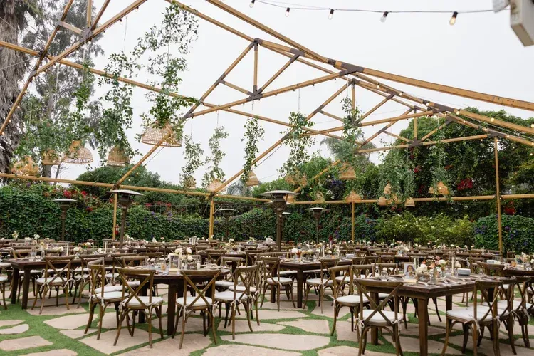 Outdoor event venue with wooden tables and chairs under a bamboo canopy, surrounded by greenery.