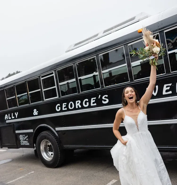Bride in white dress holding bouquet beside a black wedding bus with “George’s” on the side