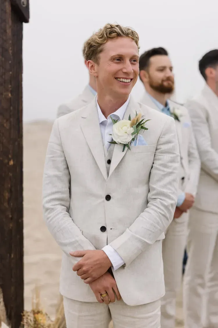 Smiling groom in a light beige suit with boutonniere, standing outdoors at a wedding ceremony