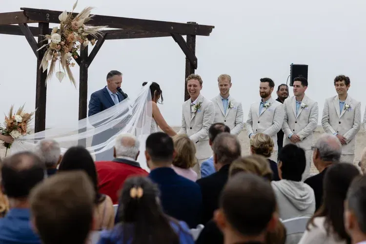 Outdoor wedding ceremony with bride and groom under wooden arch, officiant, and seated guests