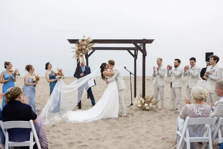 Beach wedding ceremony with bride and groom kissing under a wooden arch, guests seated nearby