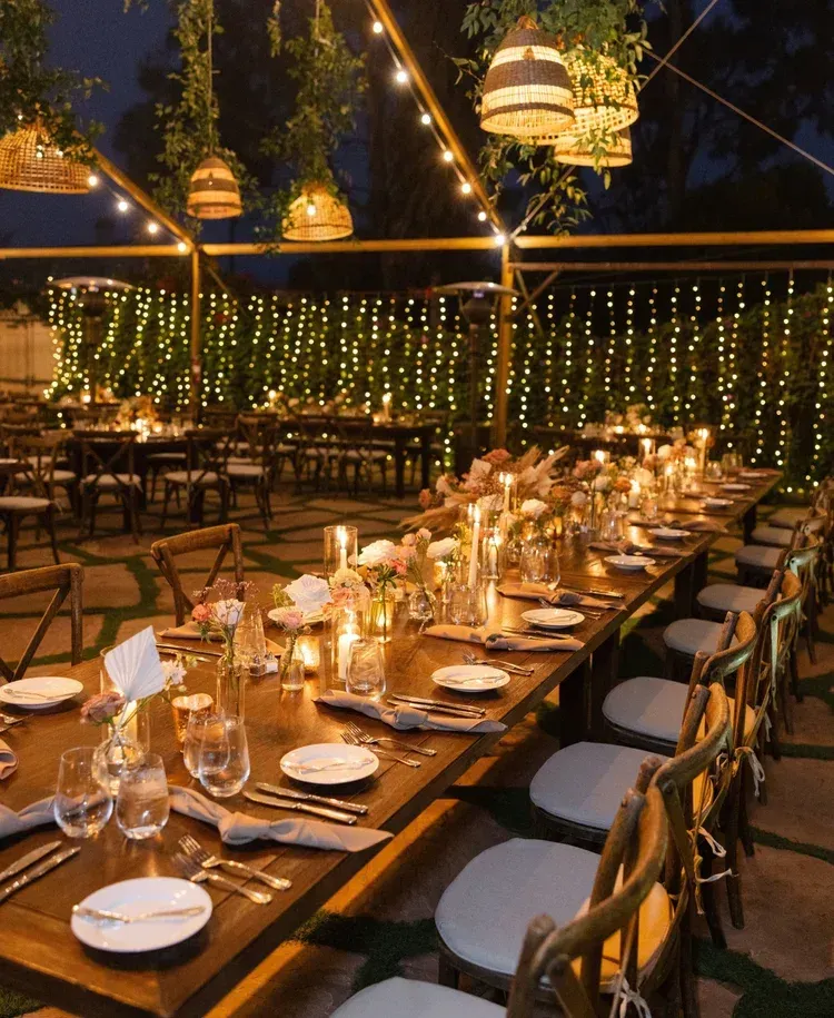 Outdoor candlelit dinner table under string lights with white plates and woven chairs at dusk