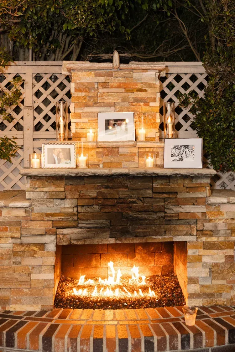 Stone outdoor fireplace with a lit fire, lattice backdrop, and framed decor on the mantel.