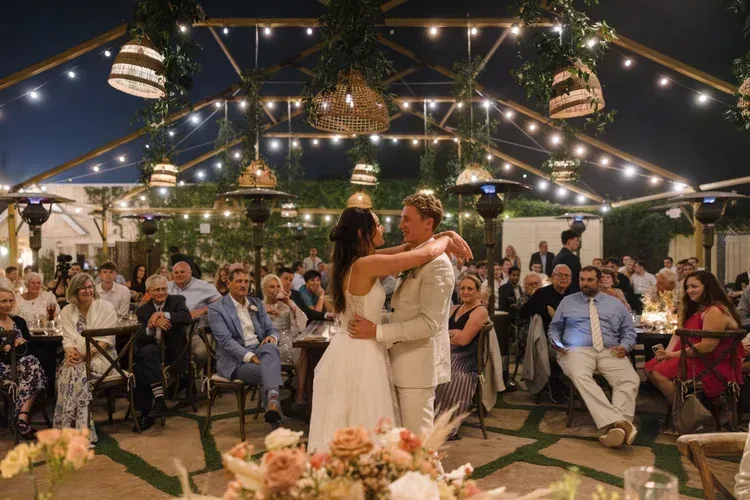 Couple embracing during a wedding reception under string lights, with seated guests around them