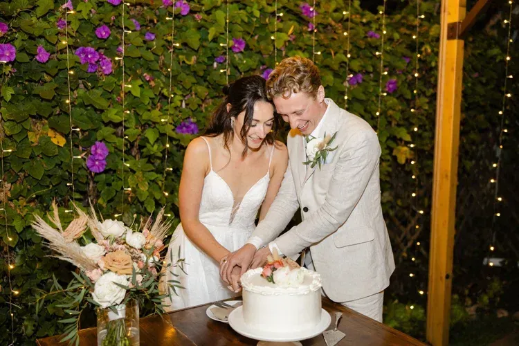 Couple in light suits cuts a white wedding cake at an outdoor reception with purple flowers behind them