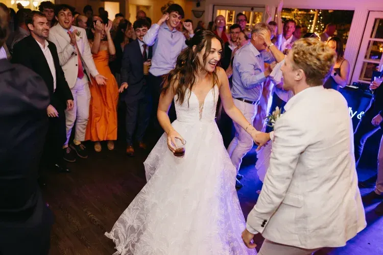 Bride and guest dancing at a wedding reception, surrounded by applauding guests and colorful lights