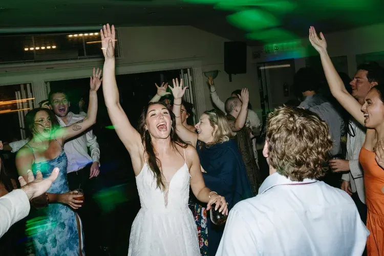 People dancing at a lively indoor party, with a woman in a white dress raising her arms.