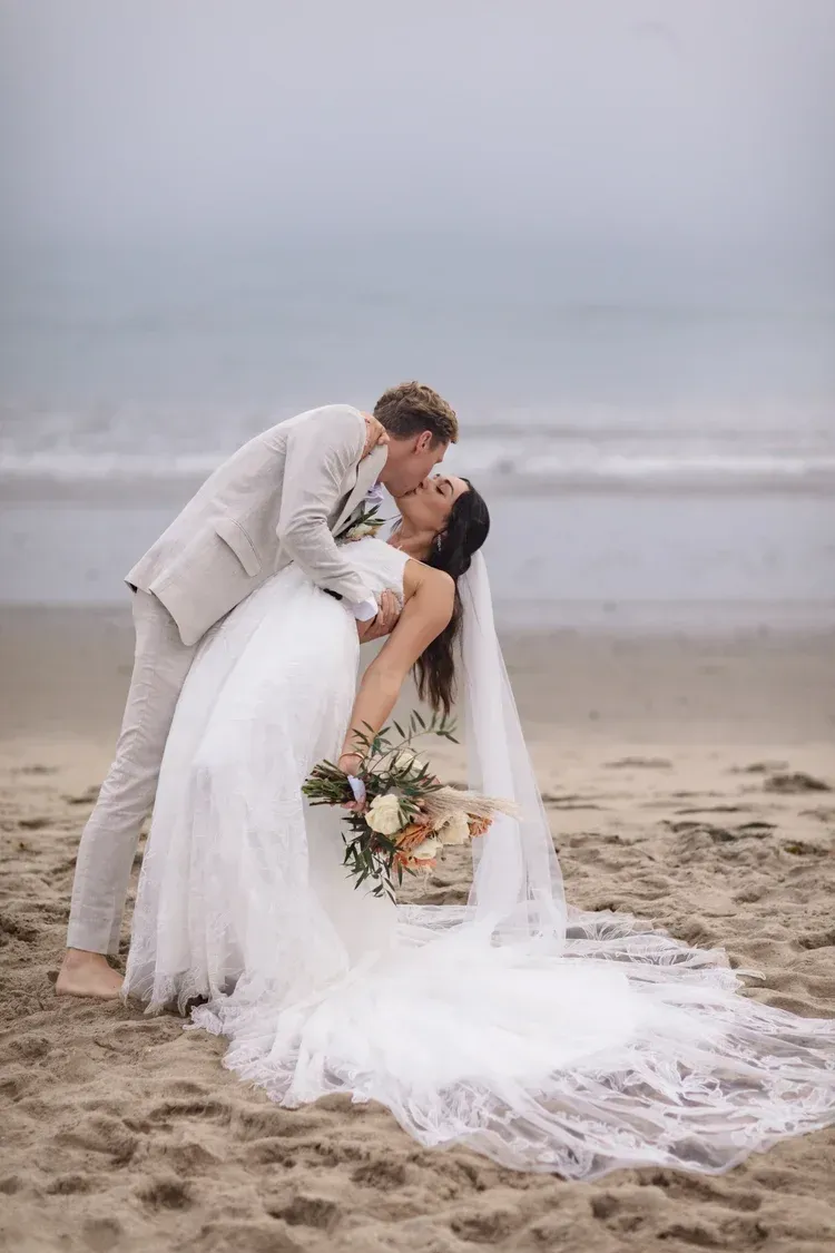Bride and groom kiss on a misty beach, bride in white dress holding bouquet.