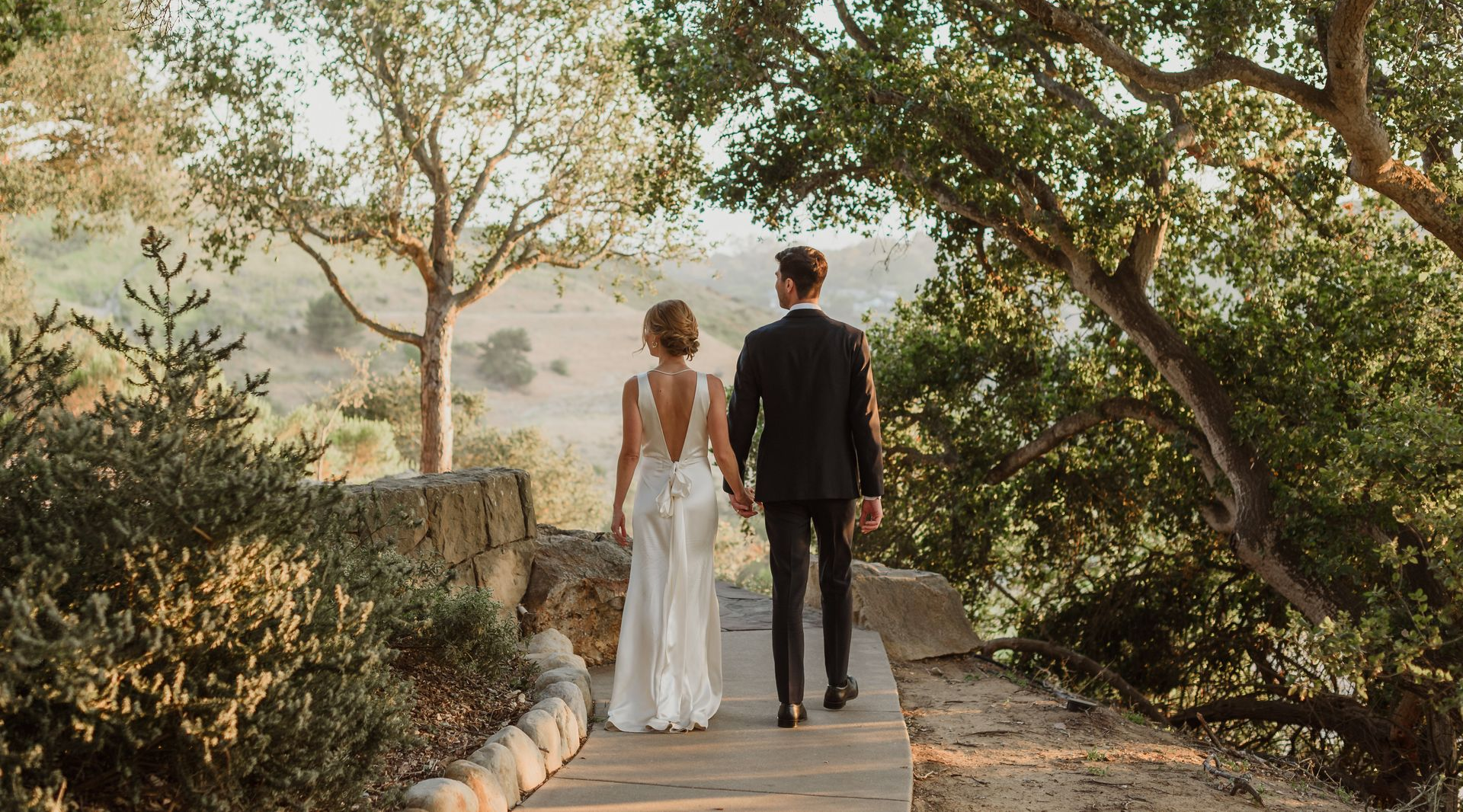Couple in wedding attire walking hand in hand down a tree-lined path outdoors