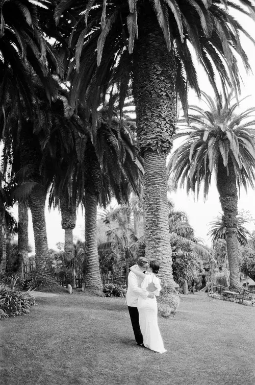 Couple in white embracing beneath tall palm trees in a garden